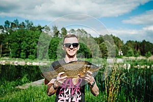 Happy cheerful young fisherman hold a big fish carp on a background of lake and nature. Fishing background. Good catch. Trophy