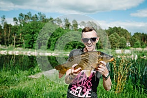 Happy cheerful young fisherman hold a big fish carp on a background of lake and nature. Fishing background. Good catch. Trophy