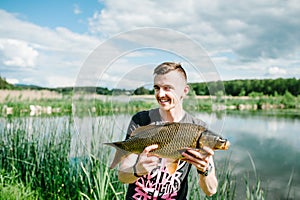 Happy cheerful young fisherman hold a big fish carp on a background of lake and nature. Fishing background. Good catch. Trophy