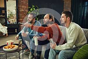 Happy and cheerful young men with alcohol drinks and snacks watching football game sitting on coach in living room at