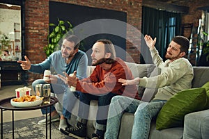 Happy and cheerful young men with alcohol drinks and snacks watching football match sitting on coach in living room at