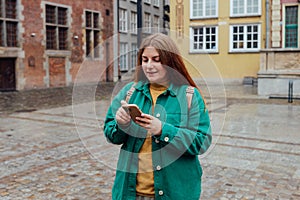 Happy cheerful young woman with backpack walking on city street checks her smartphone. Portrait of beautiful 30s girl