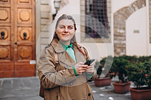 Happy cheerful young woman with backpack walking on city street checks her smartphone. Portrait of beautiful 30s girl