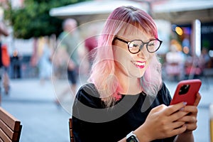 Happy cheerful young woman with multicolored pink hair wearing glasses sitting in a street eropean cafe with a smartphone in her