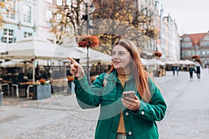 Happy cheerful young woman with phone walking on city street checks her smartphone. Redhead girl with backpack pointing