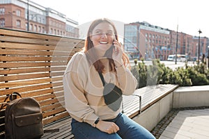 Happy cheerful young woman siting on the bench on city street, 30s Redhead girl talking on smartphone outdoors. Urban