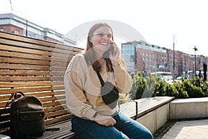 Happy cheerful young woman siting on the bench on city street, 30s Redhead girl talking on smartphone outdoors. Urban