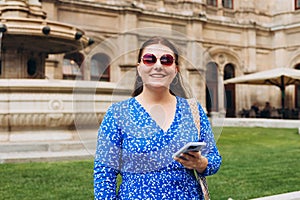 Happy cheerful young woman in sunglasses walking on city street checks her smartphone. Portrait of beautiful 30s girl