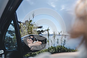 Happy cheerful young woman traveling on the sea by car. Portrait in the reflection of the rearview mirror. Summer vacation concept