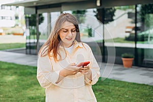 Happy cheerful young woman walking on city street checks her smartphone. Portrait of beautiful 30s businesswoman using