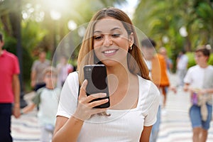 Happy cheerful young woman watching her smartphone when walking in street with blurred people on background. Millennial girl using