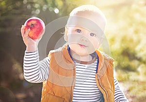 Happy child (baby boy) with red apple in the autumn (fall) day. Kid eating healthy food, snack.