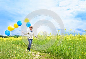 Happy child with balloons running on field.Laughing toddler boy playing in a yellow field. Blooming rapeseed, blue sky