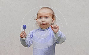 Happy child in a blue sweater eating a blue candy lollipop, on a light background. Beautiful little girl with brown eyes and a