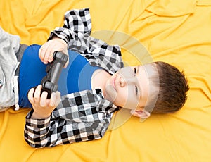 Happy child boy in checked shirt lying on the couch with black joystick in his hands playing the video game. Playing