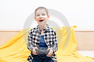 Happy child boy in checked shirt sitting on the couch with black joystick in his hands playing the video game. Playing