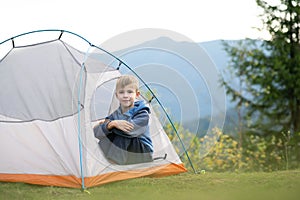 Happy child boy resting alone in a tent at mountain campsite enjoying view of beautiful summer nature. Hiking and active way of