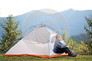 Happy child boy resting alone in a tourist tent at mountain campsite enjoying view of beautiful summer nature. Hiking and active