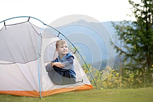 Happy child boy resting alone in a tourist tent at mountain campsite enjoying view of beautiful summer nature. Hiking and active