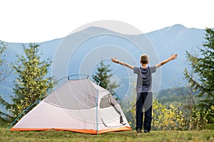 Happy child boy standing with raised hands near a tourist tent at mountain campsite enjoying view of beautiful summer nature.