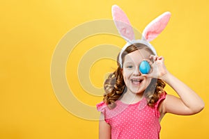 Happy child in bunny ears holds an easter blue egg. Portrait of a little girl on a yellow background