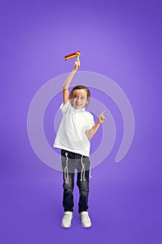 Happy child in casual attire holding colorful grogger toy and showing pointing gesture against purple studio background.