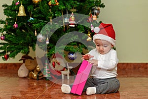 Happy child with a Christmas gift. Joyful baby at home opening a little gift while sitting in front of a Christmas tree.