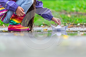 Happy child girl with colorful rubber boots playing with paper boat in puddle in autumn on nature