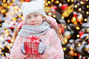 happy child girl holding christmas gift outdoor on the walk in snowy winter city decorated for new year holidays.