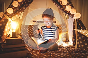 Happy child girl laughing and reading book in dark in tent at ho