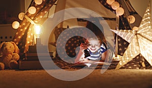 Happy child girl laughing and reading book in dark in tent at ho