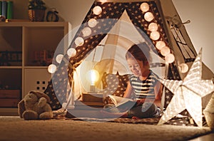 Happy child girl laughing and reading book in dark in tent at ho