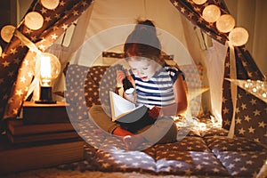 Happy child girl laughing and reading book in dark in tent at ho