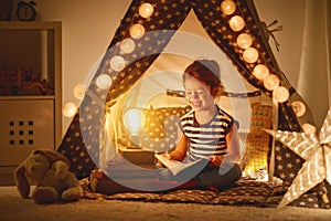 Happy child girl laughing and reading book in dark in tent at ho