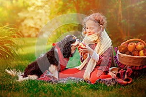 Happy child girl playing with her dog and giving him apple in sunny autumn garden