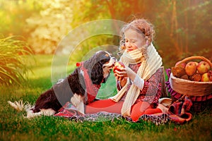 Happy child girl playing with her dog and giving him apple in sunny autumn garden photo