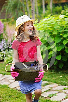 happy child girl playing little gardener and helping in summer garden, wearing hat and gloves, working with tools