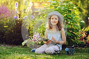Happy child girl playing little gardener in summer, wearing funny hat and holding bouquet of flowers