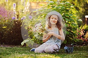 happy child girl playing little gardener in summer, wearing funny hat and holding bouquet of flowers