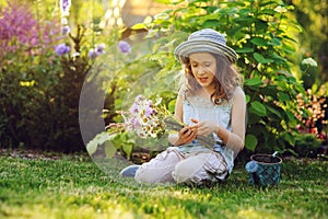 Happy child girl playing little gardener in summer, wearing funny hat and holding bouquet of flowers