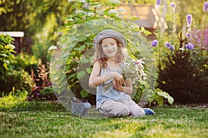 happy child girl playing little gardener in summer, wearing funny hat and holding bouquet of flowers.