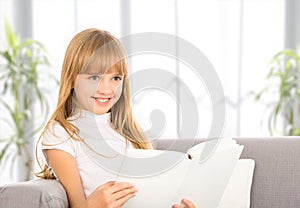 Happy child girl reading a book while sitting on sofa