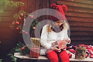 Happy child girl in red hat and scarf wrapping Christmas gifts at cozy country house, decorated for New Year and Christmas