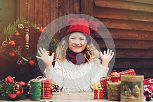 Happy child girl in red hat and scarf wrapping Christmas gifts at cozy country house, decorated for New Year and Christmas