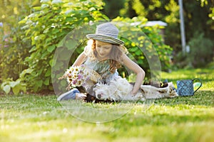 Happy child girl relaxing in summer garden with her spaniel dog, wearing gardener hat and holding bouquet of flowers