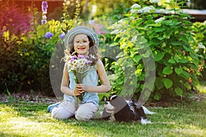 Happy child girl relaxing in summer garden with her spaniel dog, wearing gardener hat and holding bouquet of flowers