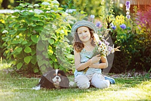 Happy child girl relaxing in summer garden with her spaniel dog, wearing gardener hat and holding bouquet of flowers.