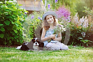 Happy child girl relaxing in summer garden with her spaniel dog, wearing gardener hat and holding bouquet of flowers