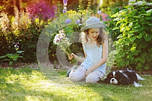 happy child girl relaxing in summer garden with her spaniel dog, wearing gardener hat and holding bouquet