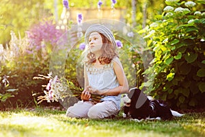 happy child girl relaxing in summer garden with her spaniel dog, wearing gardener hat and holding bouquet